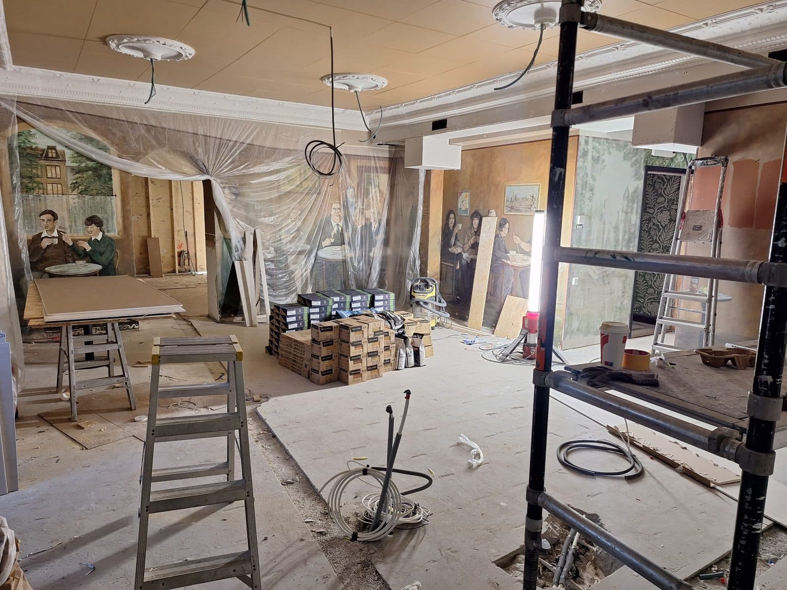 Samma kafélokal under arbetet, med byggställning, plastade väggmålningar och material på golvet
